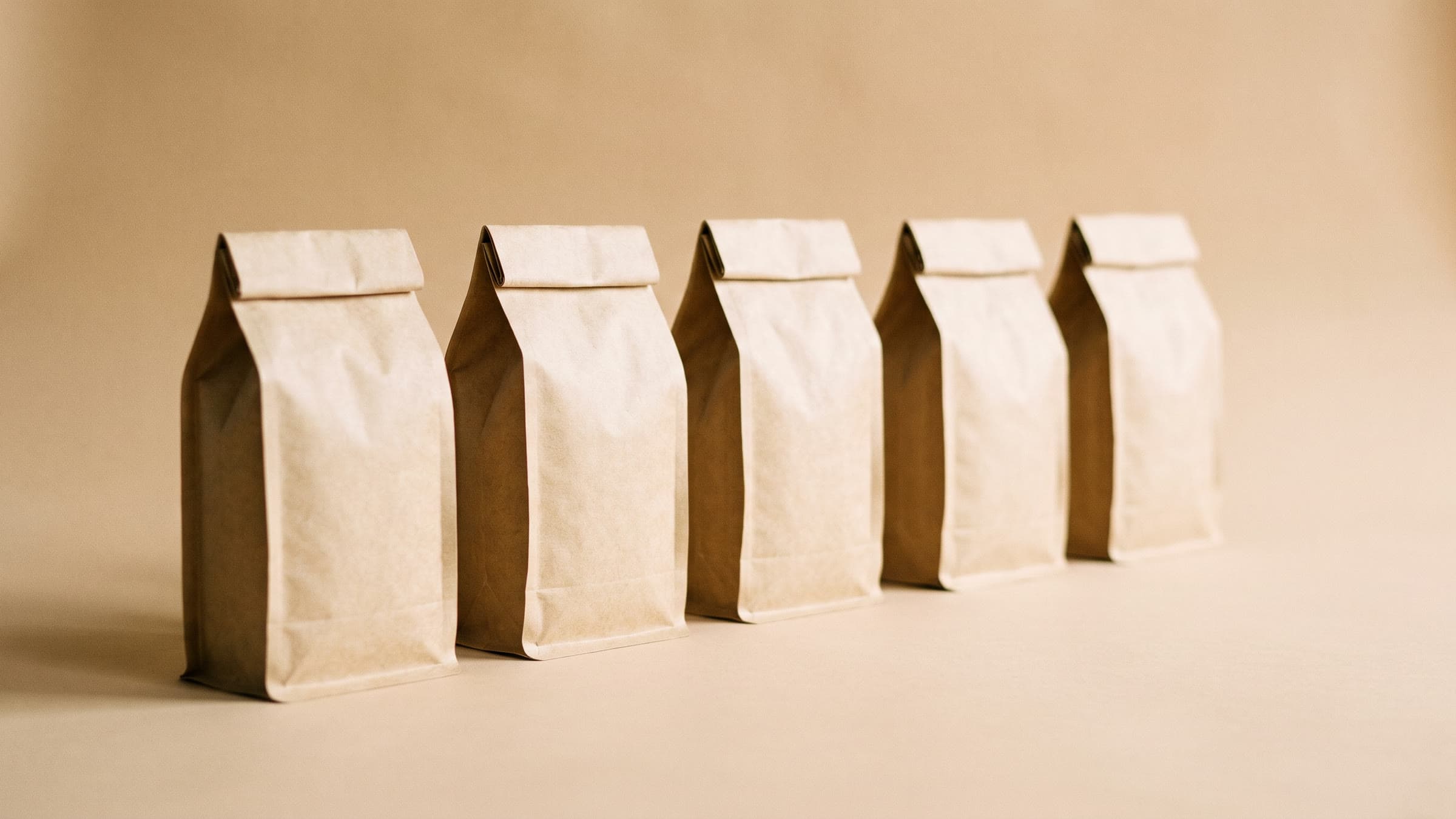 Fem sandfarvede kraftpapirsposer med kaffe på række i blødt sidelys.